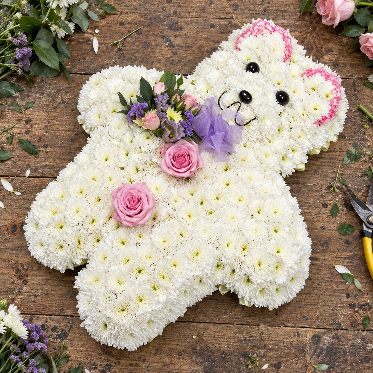 Teddy funeral tribute with white chrysanthemums, pink roses and a lilac bow on a rustic florist bench